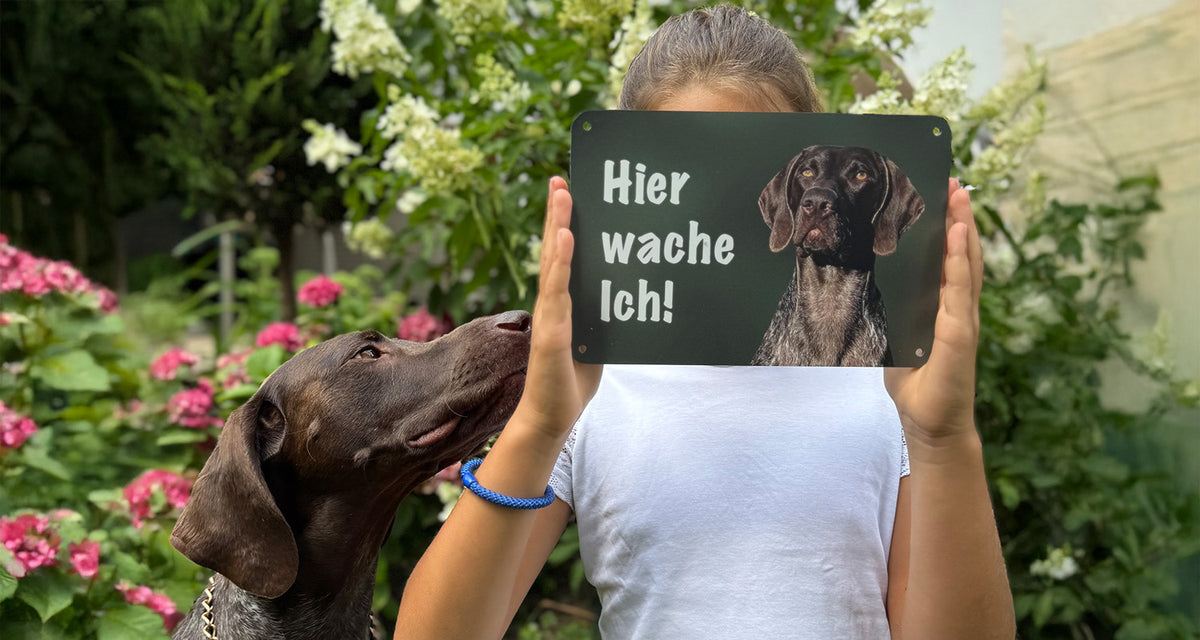 Hund neben einer Person, die ein Hundeschild mit der Aufschrift „Hier wache ich!“ hält
